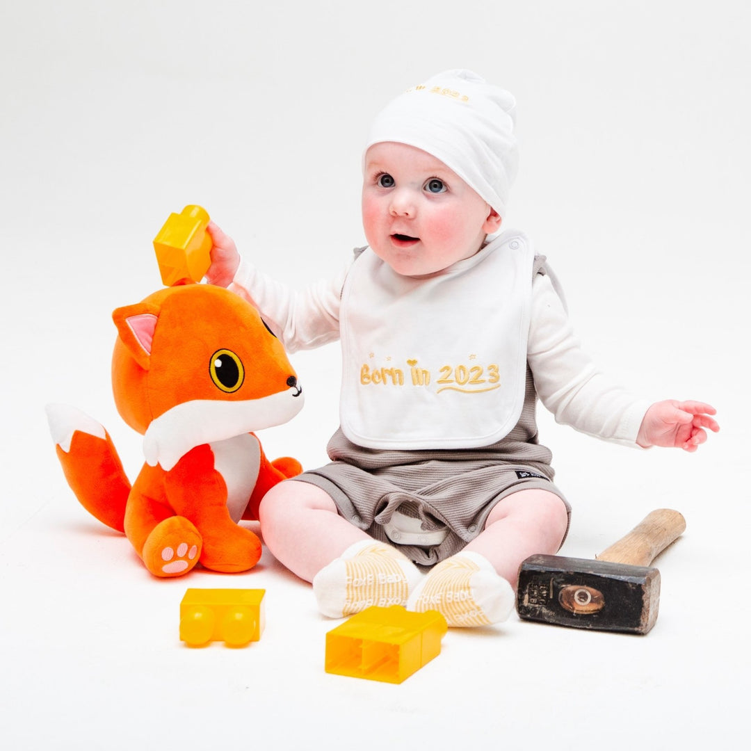 A little boy playing with a cuddly fox toy and wearing a yellow Born In 2023 baby bib, beanie, and socks set.