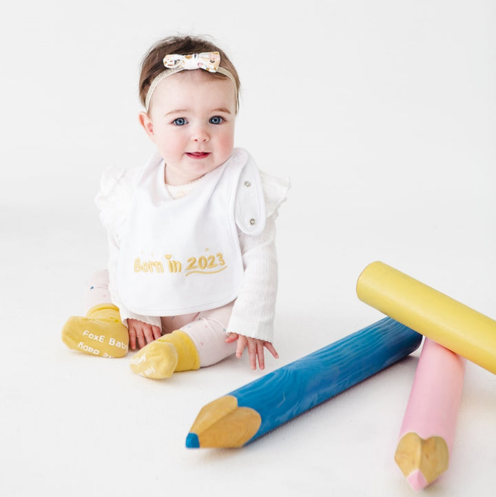 Cute baby girl wearing yellow Born In 2023 baby bib and socks, and a bow, is sitting next to oversize novelty pencils.