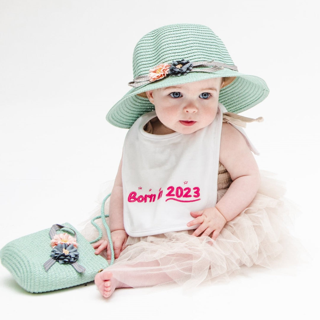 Baby girl sitting wearing a green hat and a pink Born in 2023 bbay bib.
