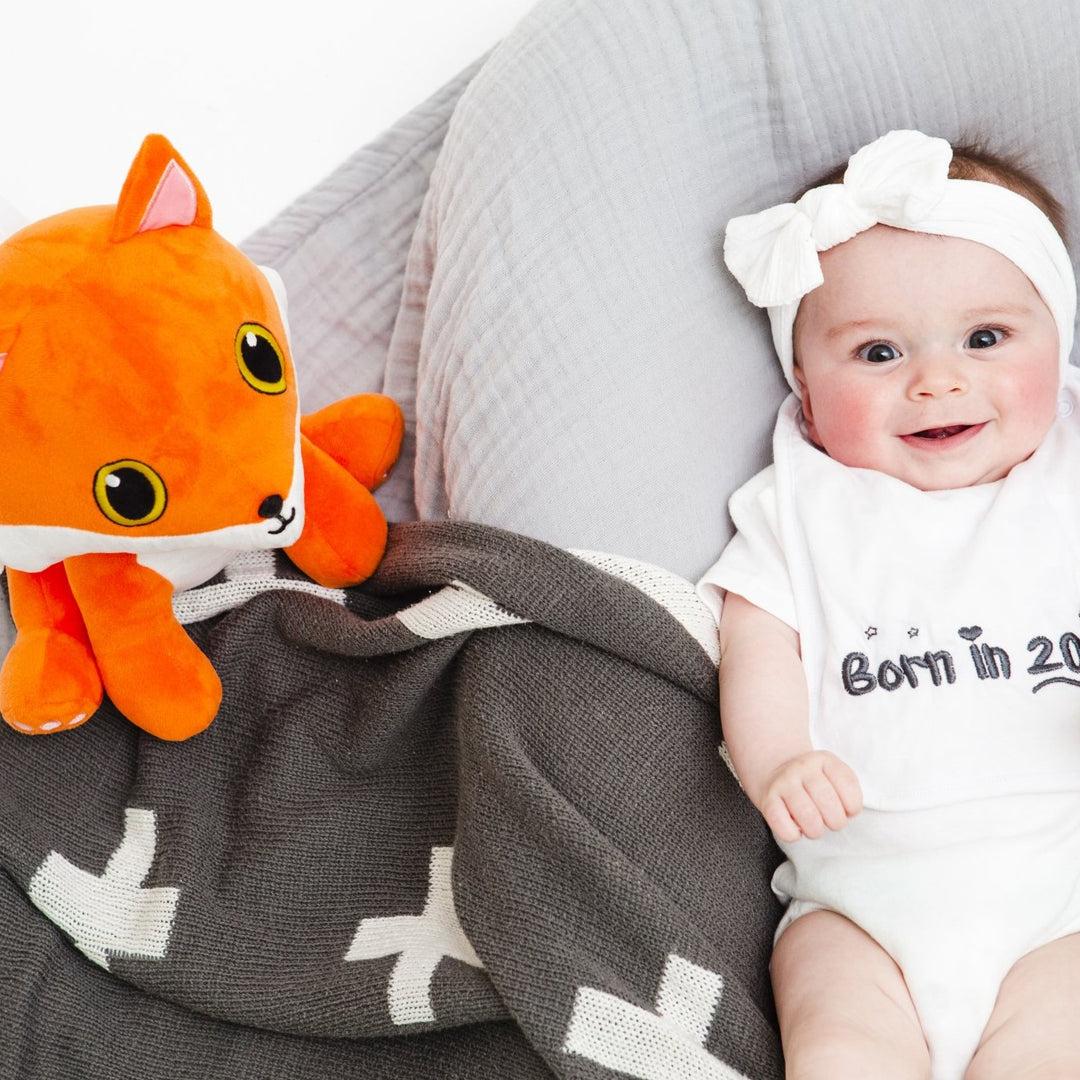 A soft toy fox sits next to a baby girl with rosie cheeks and wearing a grey Born in 2023.