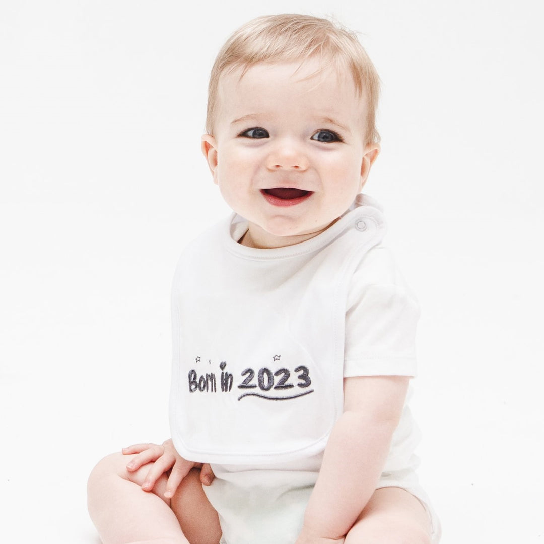 A smiling baby boy wearing a grey Born In 2023 baby beanie.