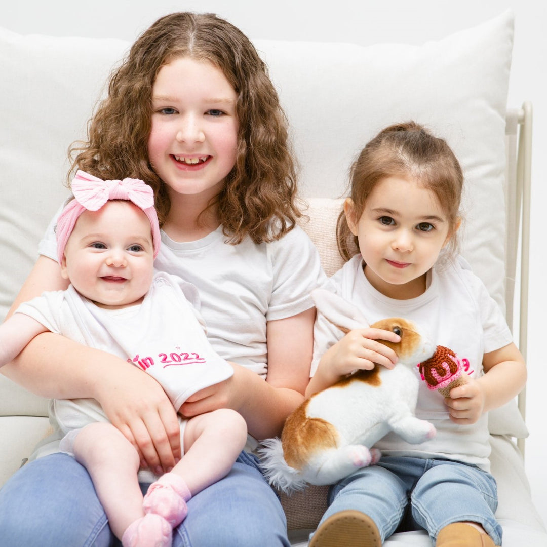 Three sisters sitting together wearing pink Born In 2023 socks and bib, one is playing with a soft toy rabbit.