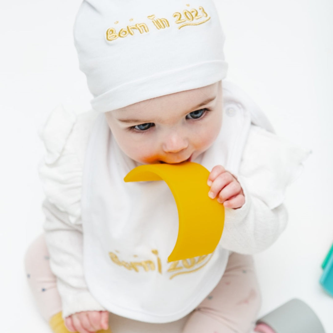 Baby girl, wearing a yellow Born in 2023 gift set including a biby bibs, beanie, and socks.
